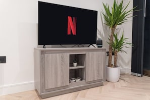 A modern living room with a flat-screen TV mounted on a wooden cabinet, a potted plant, and a small shelf with decorative items. - One bed flat 7 mins walk to Manchester Piccadilly (Greater Manchester)