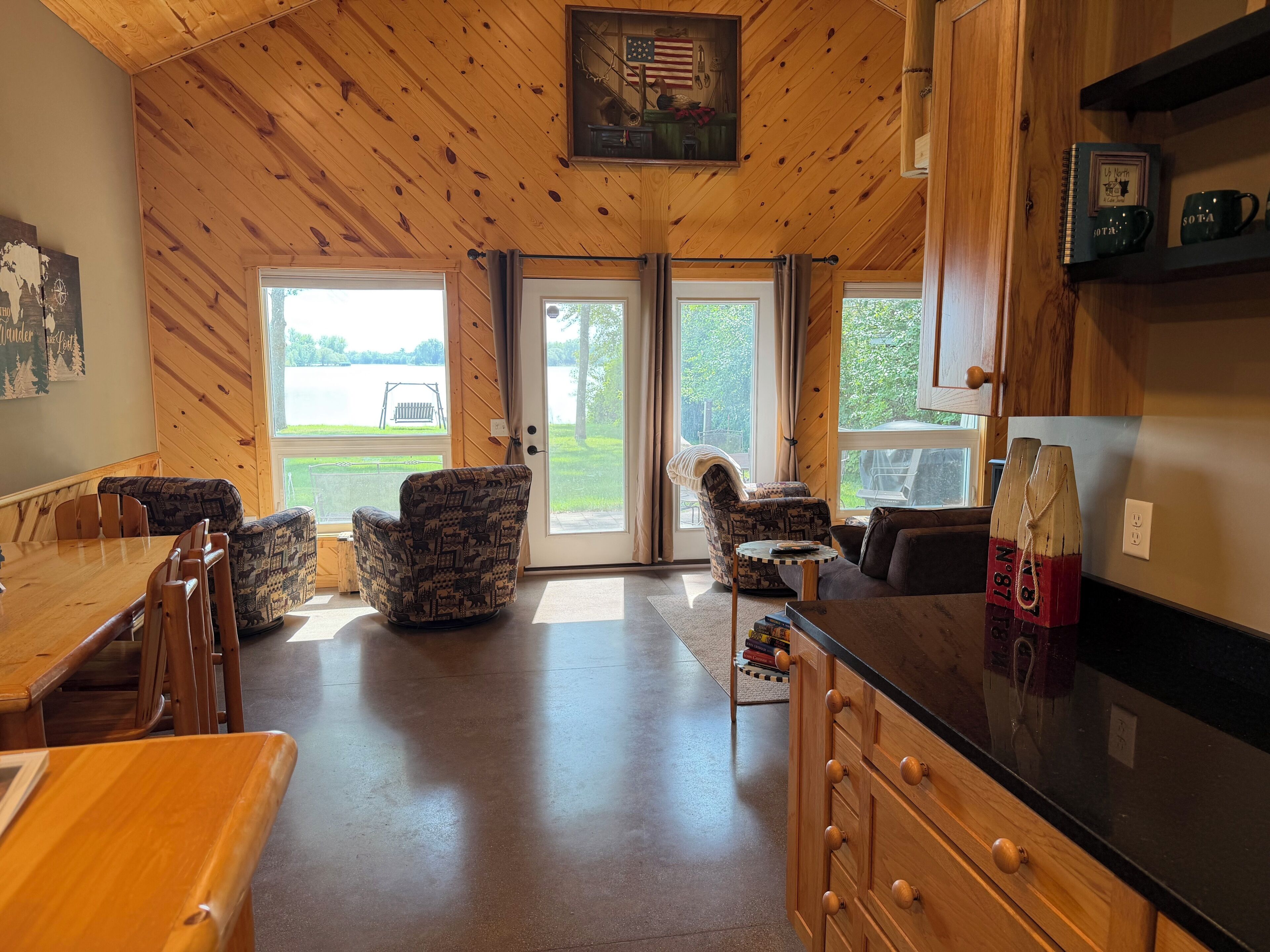 A cabin interior with a wooden ceiling, a dining area with a table and chairs, a seating area with a sofa and armchairs, and a kitchen with a refrigerator and cabinets.