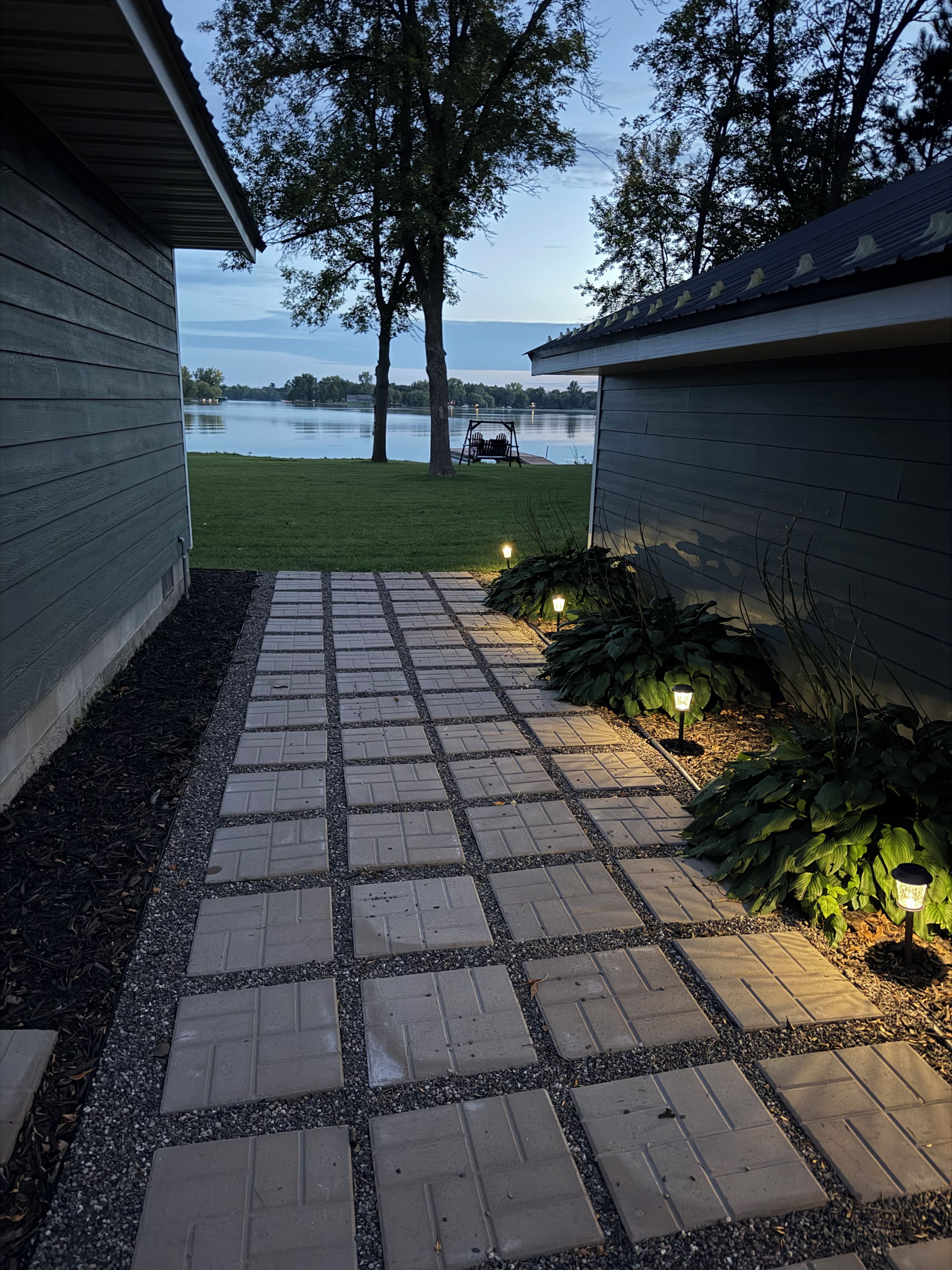 A paved pathway leading to a lake view, flanked by wooden buildings and a swing set.