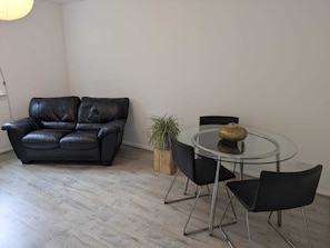 A room with a black leather sofa, a glass table with chairs, a potted plant, and a wall socket.