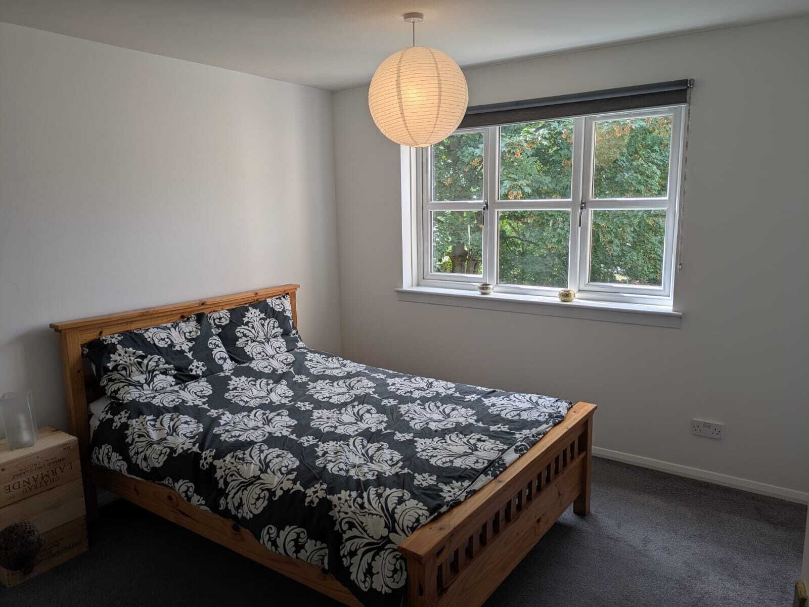 Ett sovrum med en säng i trä, ett fönster med utsikt över grönområden och en hängande lampa.