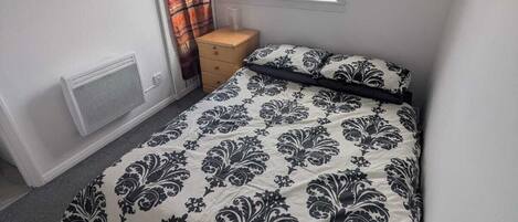 Een slaapkamer met een bed, een raam, een radiator en een klein houten nachtkastje.