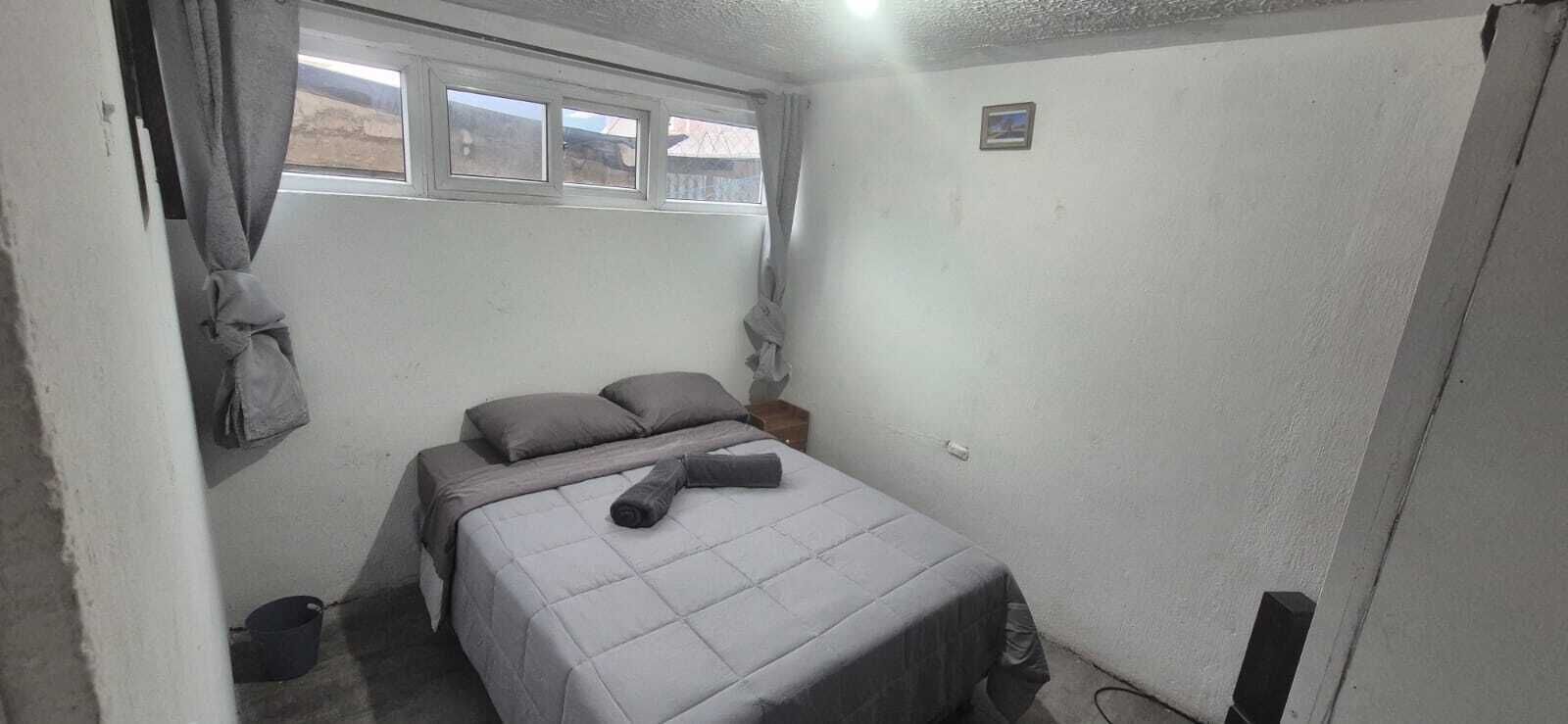 Una habitación pequeña y escasamente amoblada con una cama, un ventanal y una lámpara empotrada en la pared.