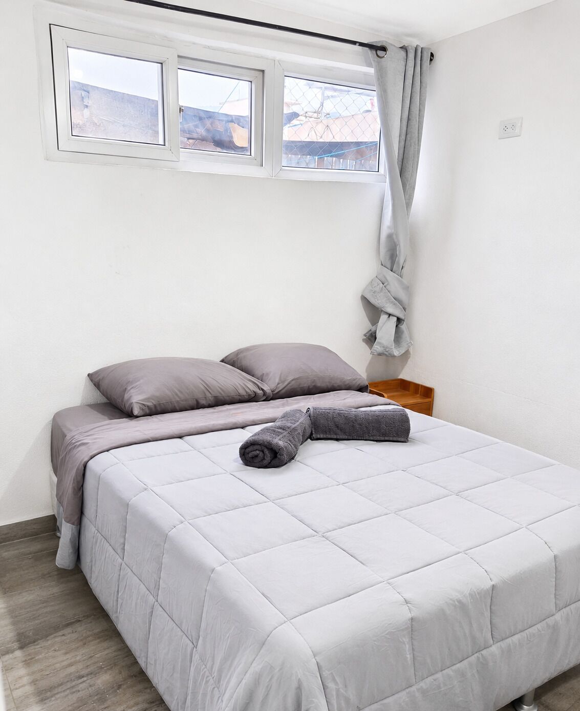 A single bed with a grey duvet and pillows, a wooden nightstand, a window with a curtain, and a view of buildings outside.