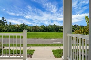 Eine Veranda mit weißen Geländern, ein gepflasterter Weg und Blick auf ein grünes Feld mit Bäumen unter blauem Himmel.