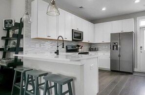 A modern kitchen with white cabinets, a stainless steel refrigerator, a microwave, and a bar area with stools. - Posh Park Circle Pad close to Charleston’s Best (North Charleston)