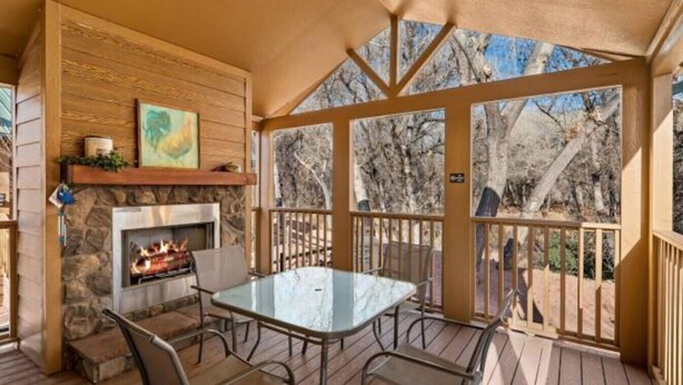 A covered outdoor area with a stone fireplace, a table with chairs, and a view of trees.
