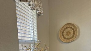 A dining area with a round table, black chairs, a vase with dried flowers, a woven wall hanging, and a window with blinds.