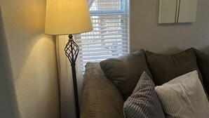 A room with a sofa, a lamp, a window with blinds, and a wall-mounted thermostat.