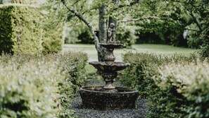 Une fontaine en pierre à plusieurs niveaux, située dans un jardin où abondent les arbustes et les arbres en arrière-plan.