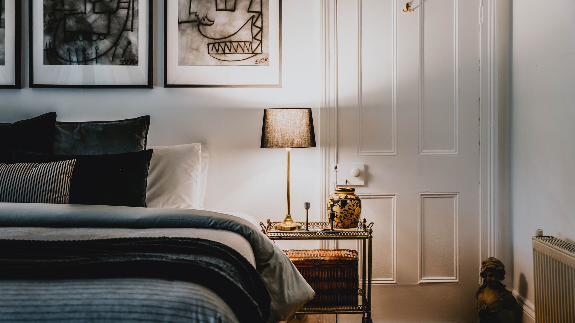 A bedroom with a bed, a bedside table with a lamp, and a wall with framed artwork.