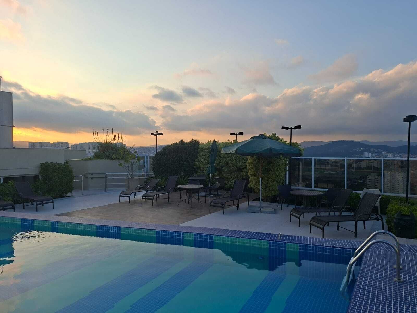 Ett poolområde på taket med solstolar, parasoller och utsikt över stadsbilden under solnedgången.
