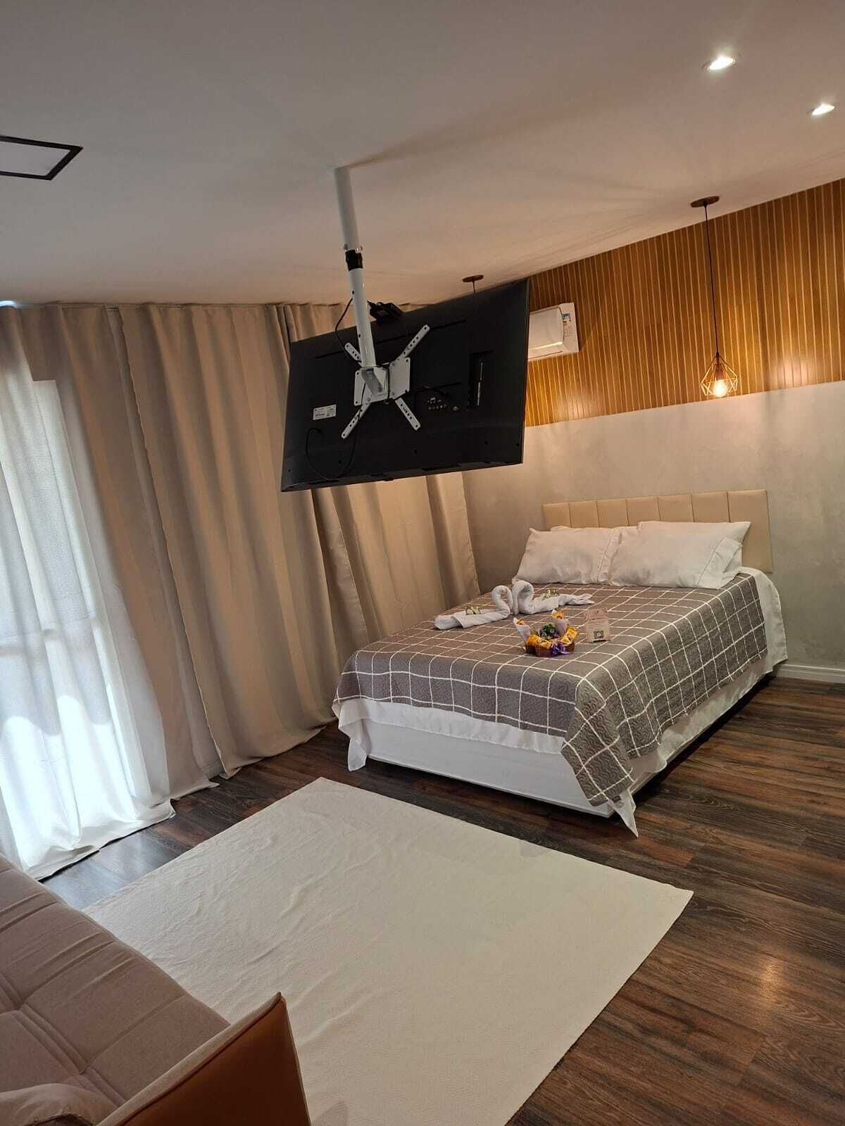 Et hotelværelse med en seng, et fladskærms-tv monteret på væggen, trægulv og et vindue med gardiner.