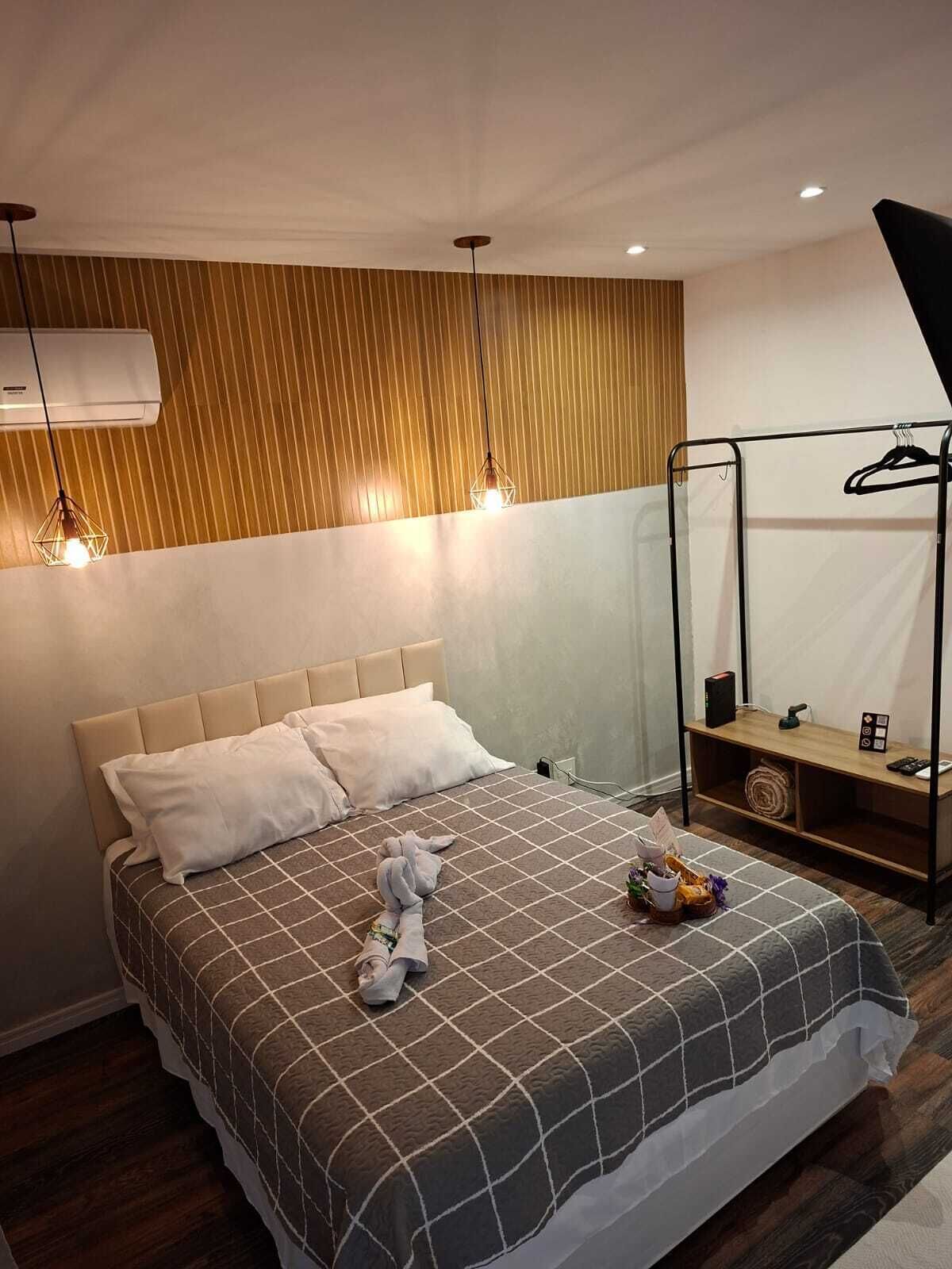 A hotel room with a bed, a hanging light fixture, a towel rack, and a small wooden shelf with items.