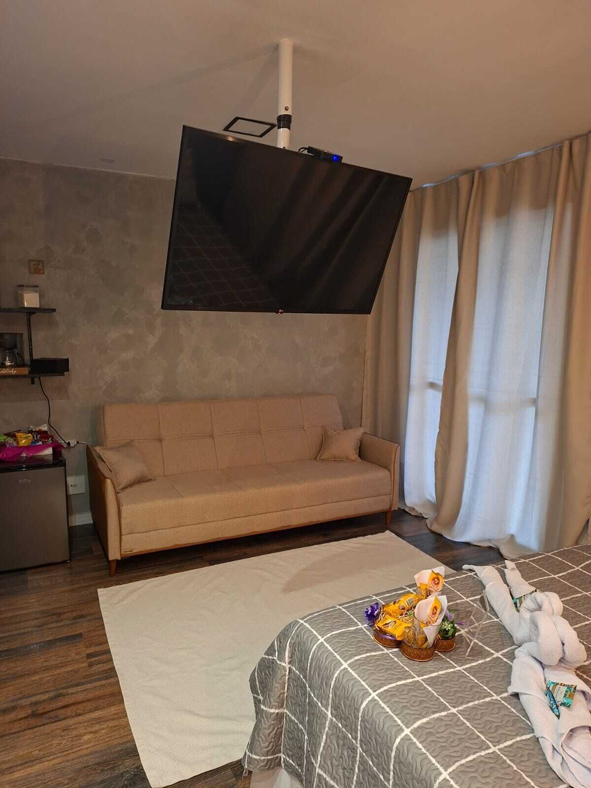 Een hotelkamer met een bank, een bed met een geruit sprei, een flatscreen televisie aan de muur en een raam met gordijnen.