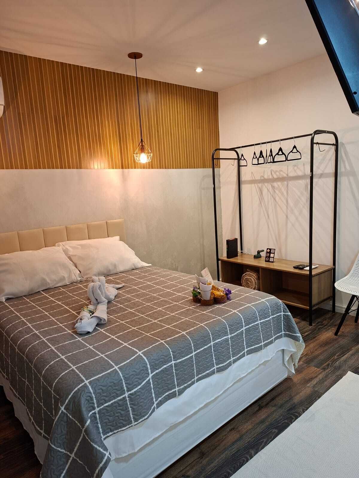 Ein ordentlich bezogenes Bett mit karierter Tagesdecke, ein Nachttisch mit Lampe und ein Kleiderständer.