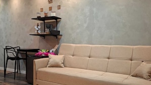 Ein modernes Wohnzimmer mit einem beigen Sofa, einem kleinen Tisch und einem Sessel. Ein Regal mit Küchenutensilien und eine Wandleuchte sind ebenfalls vorhanden.