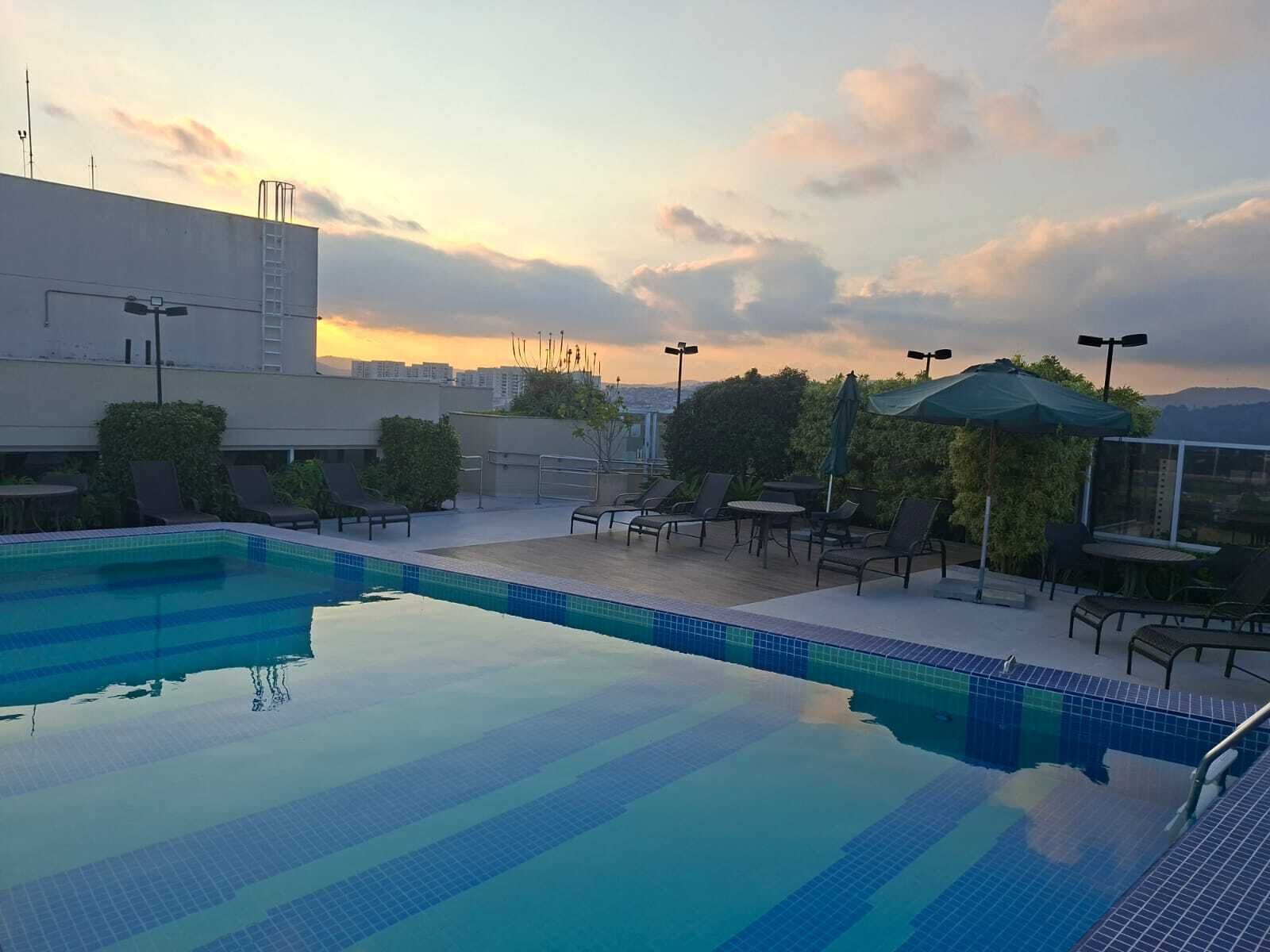 Piscina no terraço com cadeiras de descanso e guarda-sóis, cercada por edifícios e vista para o pôr do sol.