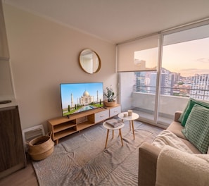 Ein modernes Wohnzimmer mit einem Flachbildfernseher, einem runden Spiegel, einer Couch, einem kleinen Tisch und Blick auf eine Stadtlandschaft durch große Fenster.