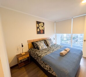 Ein Schlafzimmer mit einem Bett, einem Nachttisch, einer Wandlampe und einem Fenster mit Blick auf Gebäude.