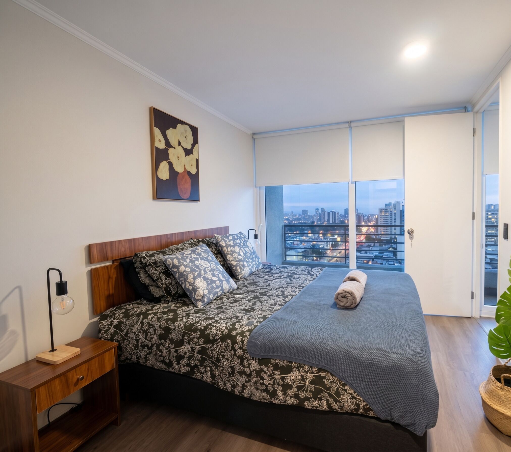A bedroom with a city view, a bed with patterned bedding, a bedside lamp, and a painting on the wall.