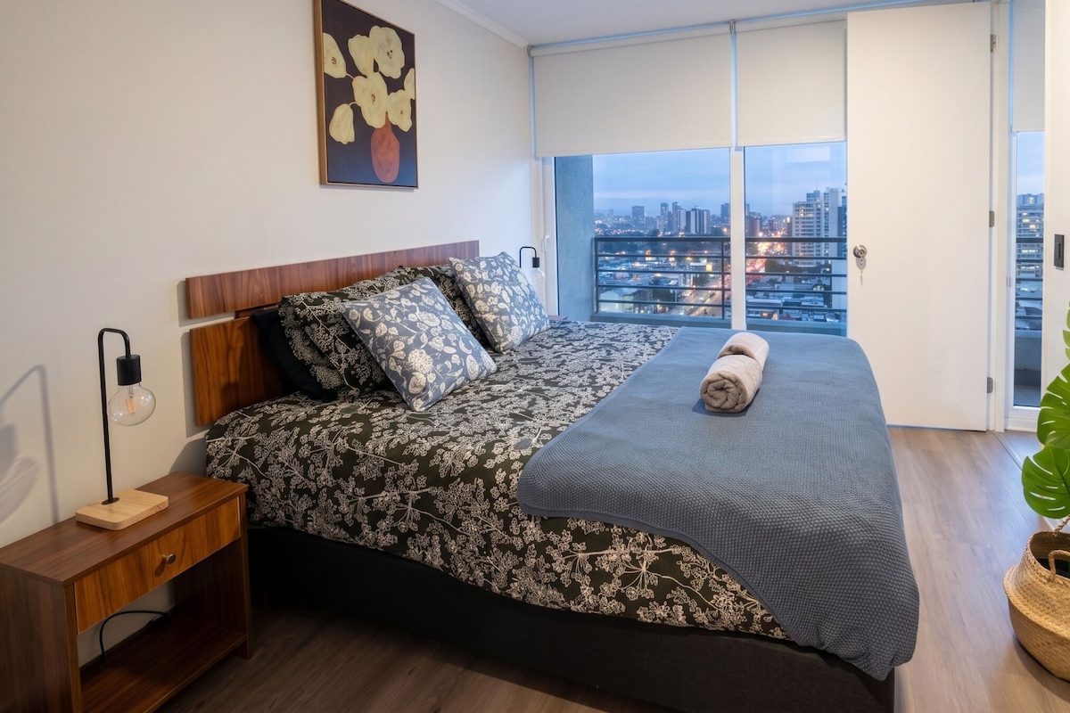 Un dormitorio con vista a la ciudad, una cama con ropa de cama estampada, una lámpara de noche y un cuadro en la pared.