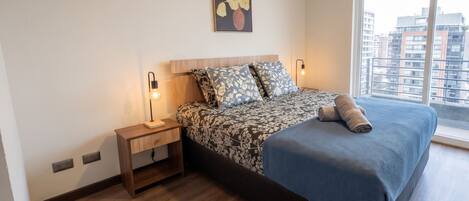 A bedroom with a bed, bedside tables, a night lamp, a wall-mounted artwork, and a large window with a view of buildings.