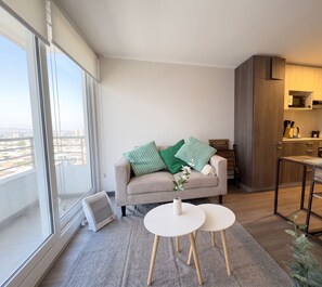 A modern living room with a sofa, two small tables, a view of the city, and a kitchen area.