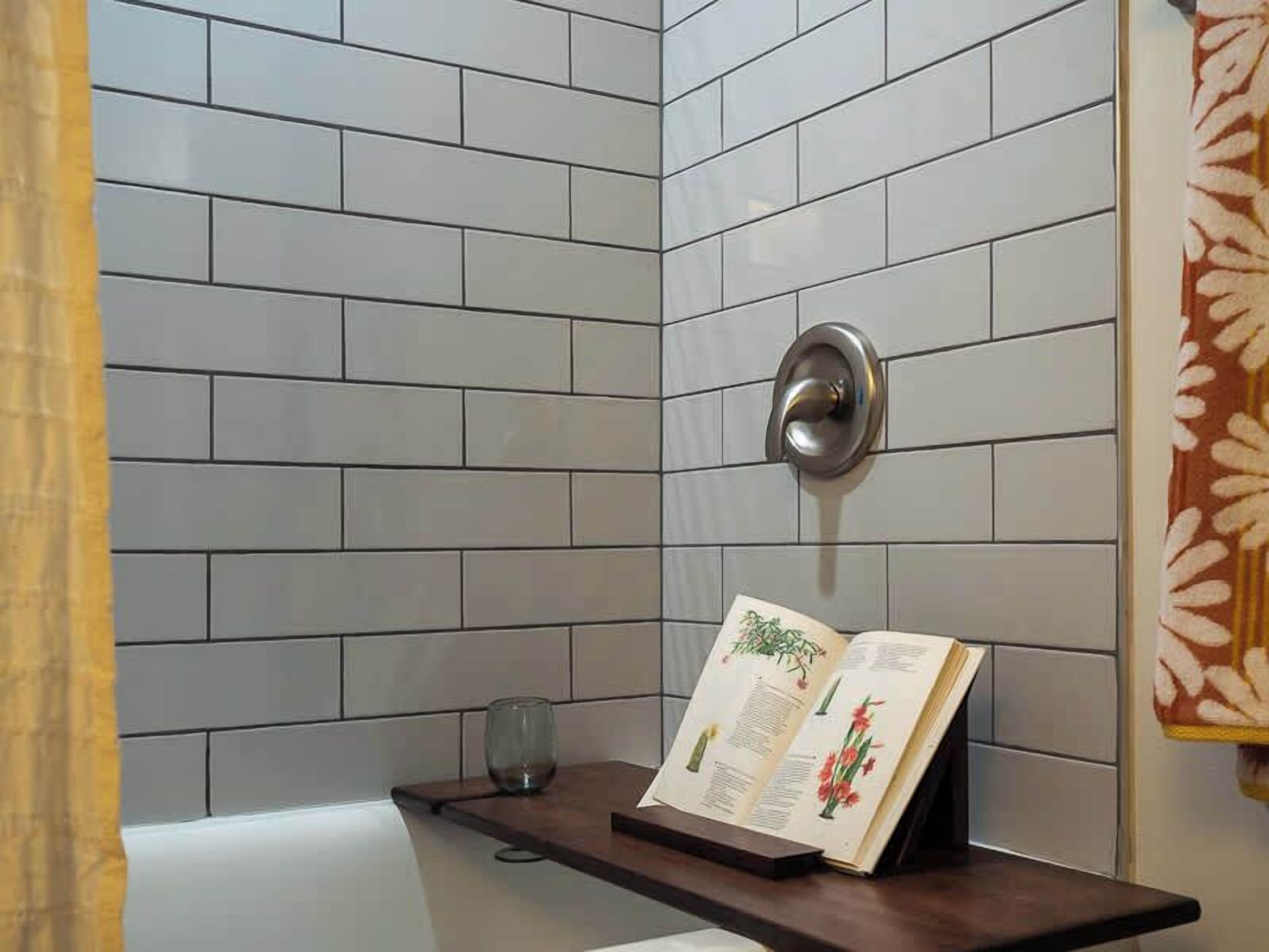 Baño en esquina con paredes de azulejos blancos, un estante de madera con un libro, un vaso y un perchero.