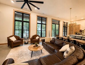 A spacious living room with large windows, a ceiling fan, and a dining area with wooden cabinets. - The Shipwreck Shack - Fall Getaway | Hot Tub • Sleeps 18 (Mauston)