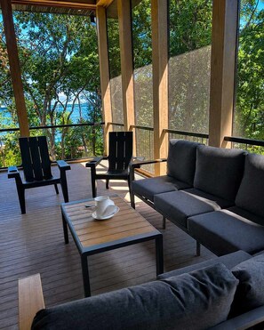 A covered outdoor seating area with a sofa and chairs, a table with a cup and saucer, and a view of trees and water. - The Shipwreck Shack - Fall Getaway | Hot Tub • Sleeps 18 (Mauston)
