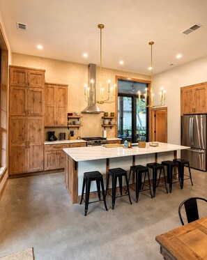 A modern kitchen with wooden cabinets, a large island with stools, stainless steel appliances, and pendant lighting. - The Shipwreck Shack - Fall Getaway | Hot Tub • Sleeps 18 (Mauston)