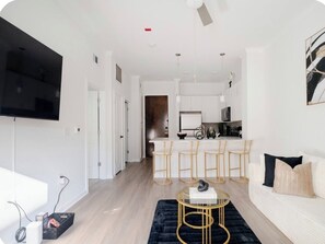 Una sala moderna con un sofá, una mesa de centro y un televisor. Se observa una zona de cocina con una barra y taburetes.
