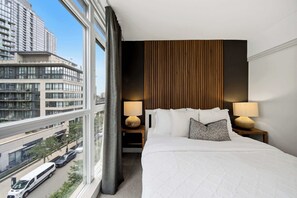 A hotel room with a large bed, two bedside lamps, a view of a cityscape, and a window with a curtain. - DT Gem near the CN Tower & Rogers Centre! (Toronto)