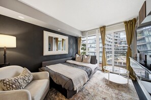A modern hotel room with a large bed, a sofa, a small table, and a view of the cityscape through large windows. - DT Gem near the CN Tower & Rogers Centre! (Toronto)