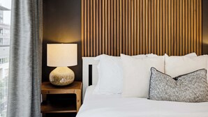 Ett hotellrum med en säng, ett nattduksbord med en lampa och utsikt över staden genom ett fönster.