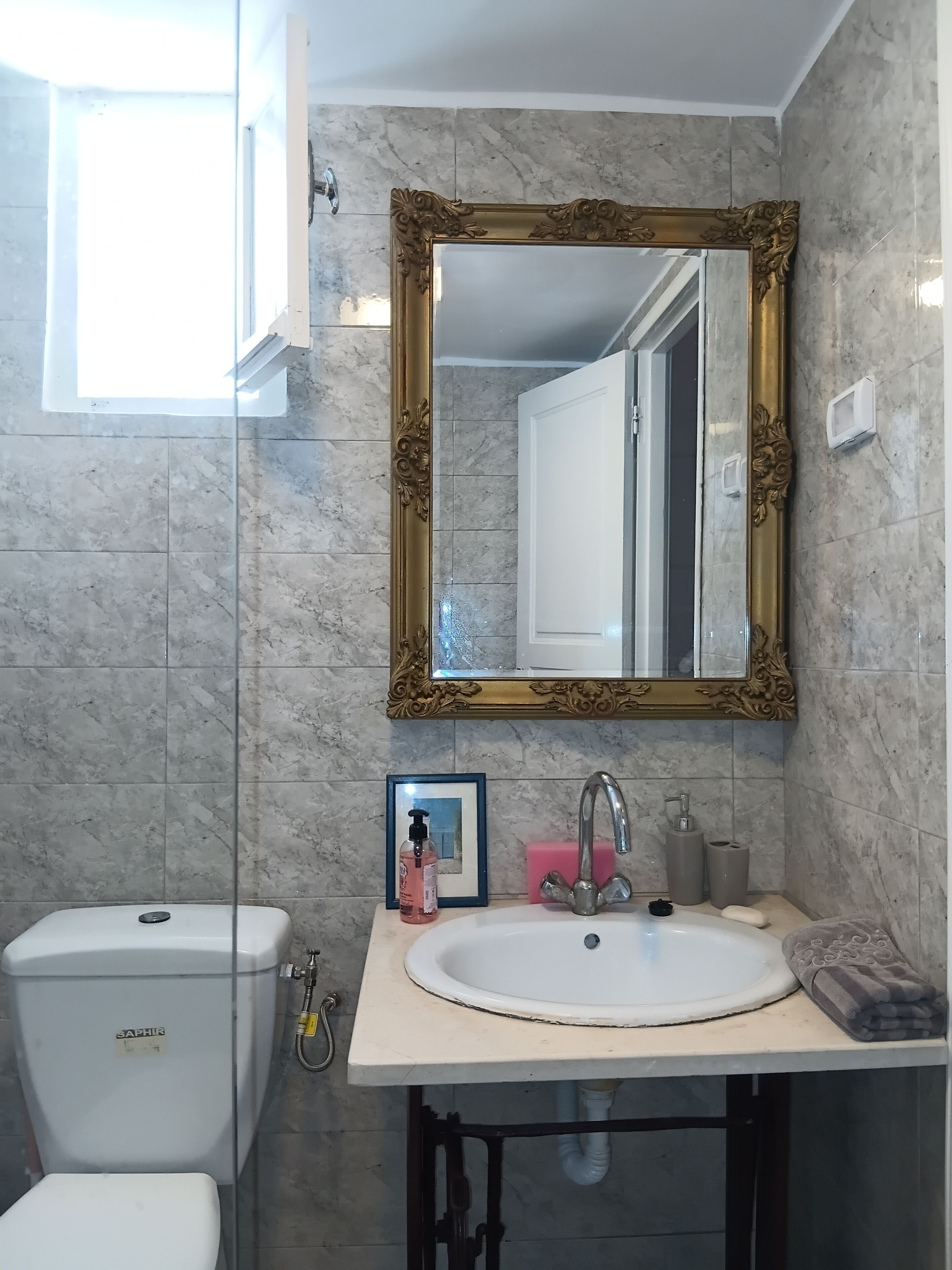 Un baño con pared de mármol, un espejo con marco dorado, un lavamanos blanco, un inodoro y un cuadro enmarcado.