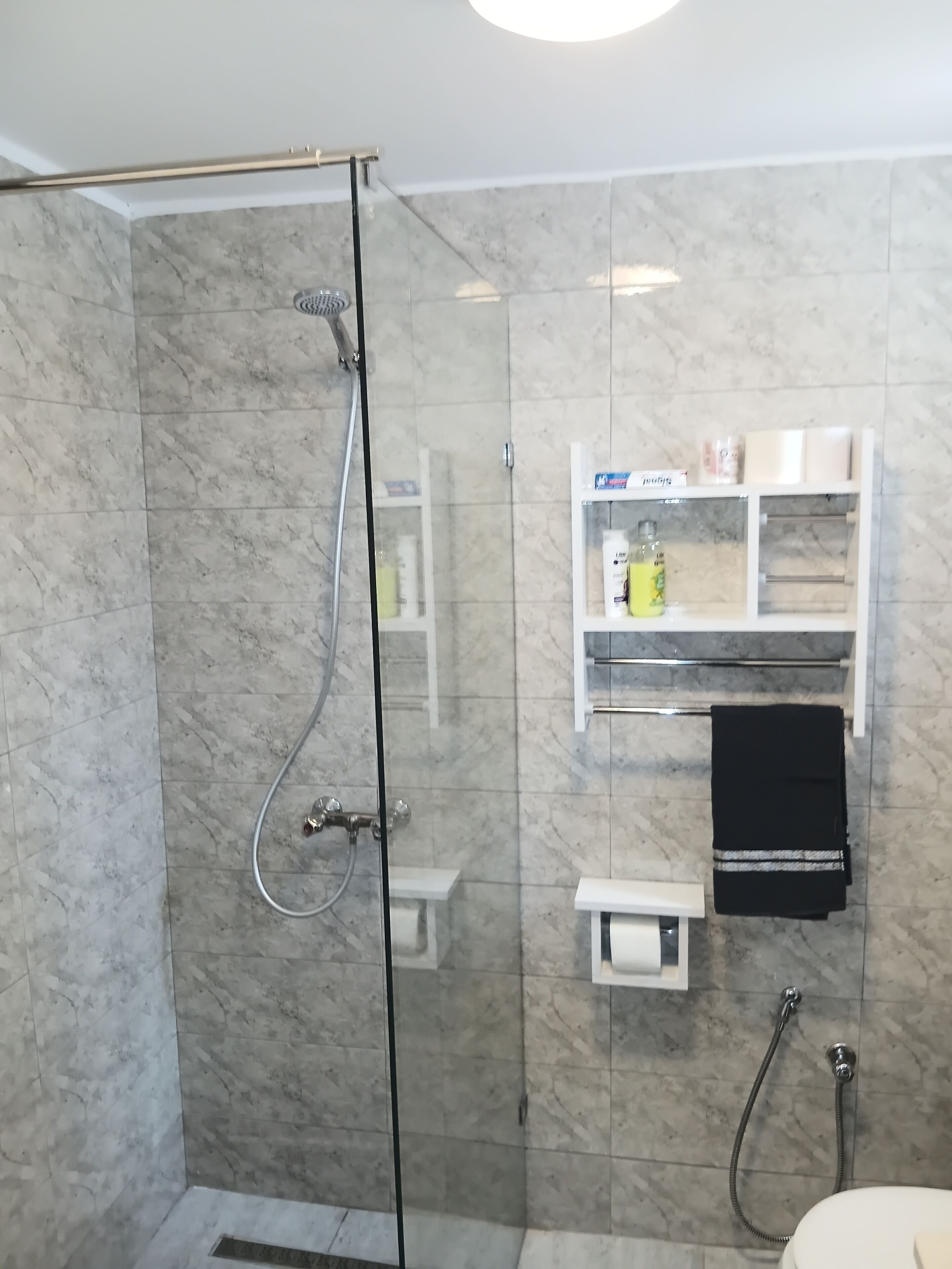 Een douchecabine met glazen wanden, een handdouche, een handdoekrek en een plank met toiletartikelen.