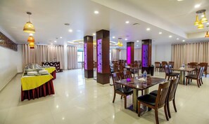 A spacious dining area with multiple tables and chairs, a buffet setup on the left, and decorative lighting fixtures. - Treebo Tryst Palmyra Milford (Ambalapuzha)