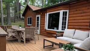 A wooden cabin with a deck, furnished with a sofa, tables, and chairs. - Cozy cabin on All Sports Lake with private beach! Close to ski resorts & trails (Atlanta)