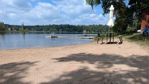 A sandy beach with a bench and a picnic table, a small boat docked, and a forested shoreline in the background. - Cozy cabin on All Sports Lake with private beach! Close to ski resorts & trails (Atlanta)