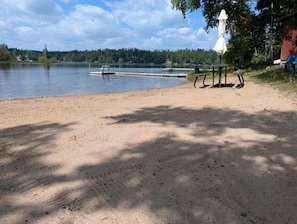 A sandy beach with a bench and a picnic table, a small boat docked, and a forested shoreline in the background. - Cozy cabin on All Sports Lake with private beach! Close to ski resorts & trails (Atlanta)