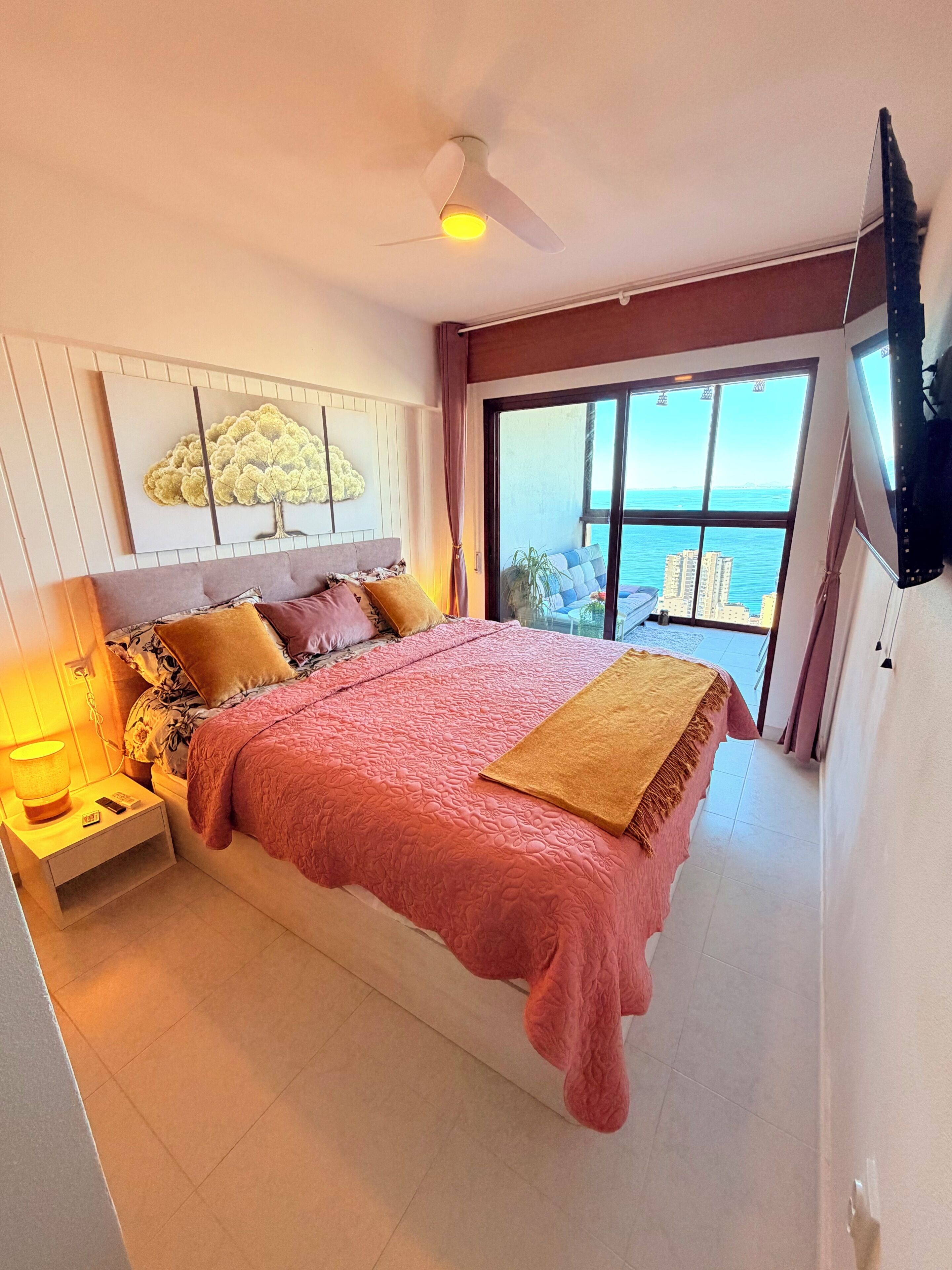A bedroom with a large bed, a ceiling fan, a nightstand, a lamp, and a view of the sea through a large window.