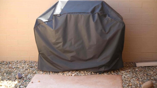 Un barbecue, recouvert d’une bâche de protection, est posé sur une surface de gravier, adossé à un mur de briques.