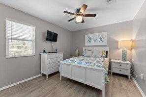 Ein Schlafzimmer mit einem Bett, einem Nachttisch, einer Kommode, einem Deckenventilator, einem Fernseher und einem Fenster mit Jalousien.