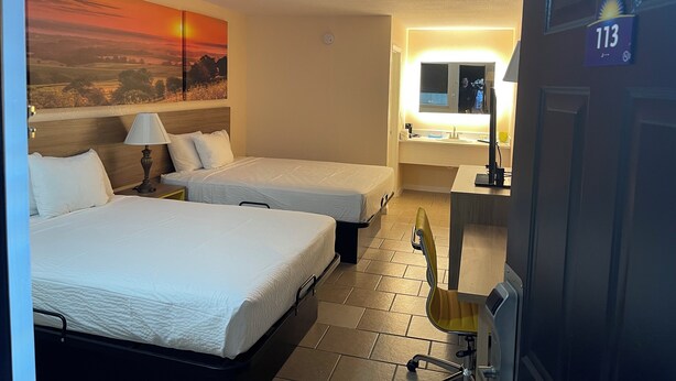 Habitación de hotel con dos camas, un cuadro de un atardecer, un baño con espejo y una puerta con la etiqueta '113'.