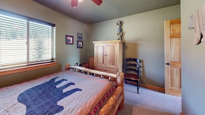 A bedroom with a bed, a wooden cabinet, a chair, and a door. - Master ensuite | Wood burning fireplace | Bike & Ski Gear Storage | Byrd's Nest (Angel Fire)