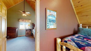 A bedroom with a wooden ceiling, a bed with a quilt, a painting of a snowy landscape, and a wooden dresser. - Master ensuite | Wood burning fireplace | Bike & Ski Gear Storage | Byrd's Nest (Angel Fire)