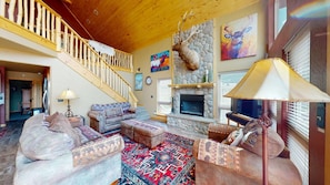 A spacious living room with a stone fireplace, a wooden staircase, and a mounted elk head. - Master ensuite | Wood burning fireplace | Bike & Ski Gear Storage | Byrd's Nest (Angel Fire)