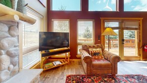 A living room with a television, a sofa with decorative pillows, a stone wall, and a wooden floor. - Master ensuite | Wood burning fireplace | Bike & Ski Gear Storage | Byrd's Nest (Angel Fire)